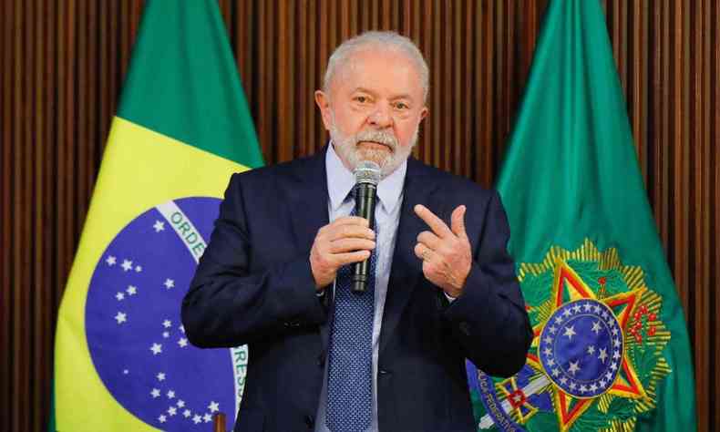 Lula falando ao microfone. Ele  um homem branco de cabelos brancos e usa terno. No fundo duas bandeiras, uma do Brasil e uma da presidncia