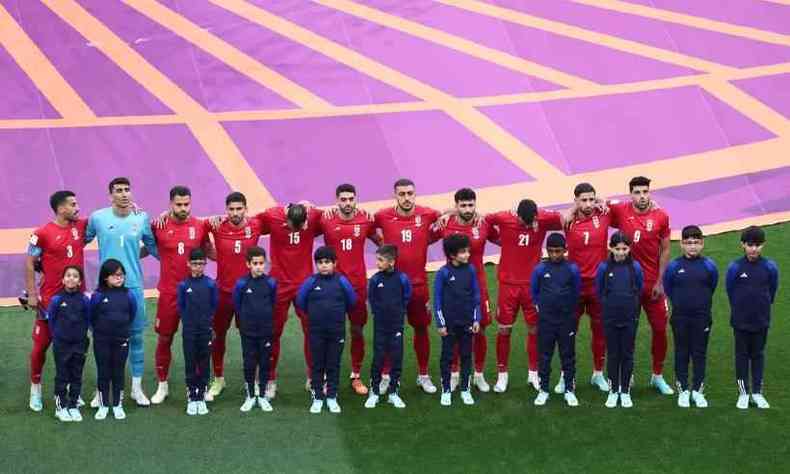 Jogadores da seleo de futebol iraniana esto alinhados em uma fila lado a lado com crianas em sua frente. Eles usam um uniforme vermelho e as crianas, azul