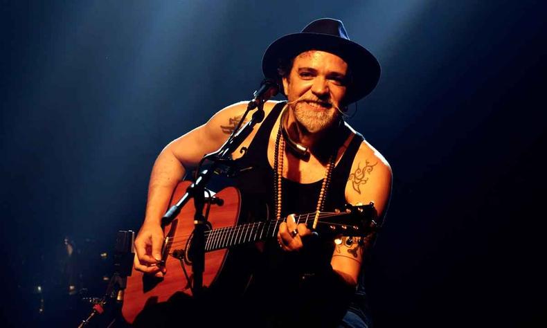 Paulinho Moska de camiseta e chapu pretos, tocando violo, Paulinho Moska sorri