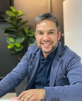 Homem branco com cabelos grizalhos sentado com blazer azul sorrindo para a foto