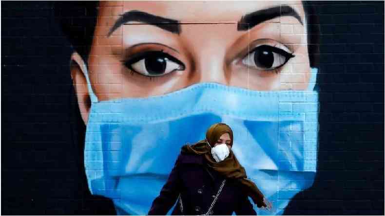 Mulher diante de mural em que aparece uma mulher de máscara