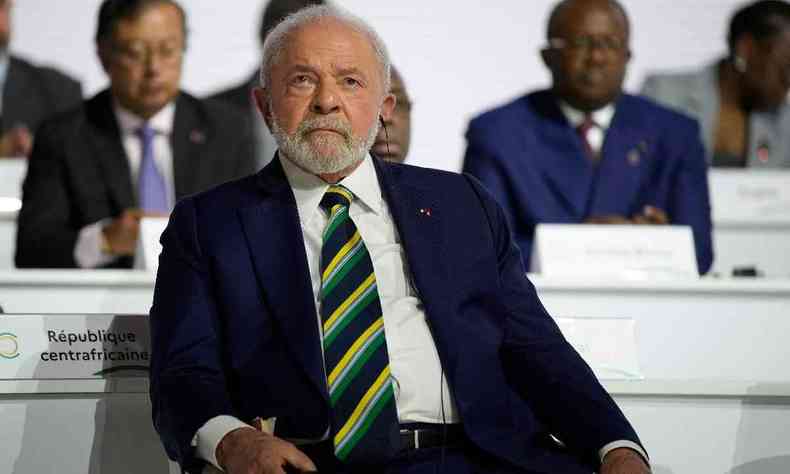 Presidente Lula sentado. No fundo, dois homens aparecem em segundo plano. Lula  um homem branco de cabelos e barba branca. Usa um terno e uma gravata listrada