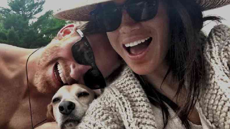 Selfie colorida mostra um casal rindo com um cachorro entre eles
