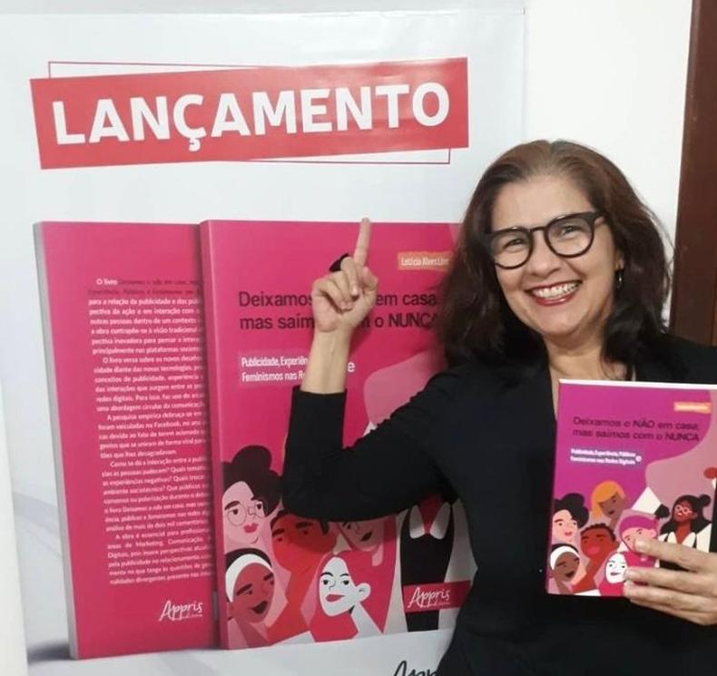 Pesquisadora Letcia Lins usa culos, segura um livro e aponta para o cartaz de lanamento da obra