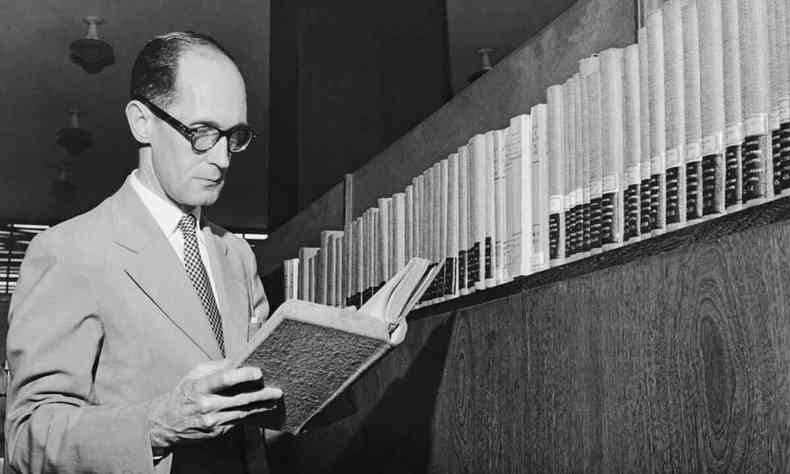 Carlos Drummond de Andrade l um livro na frente de estante com prateleira lotadas