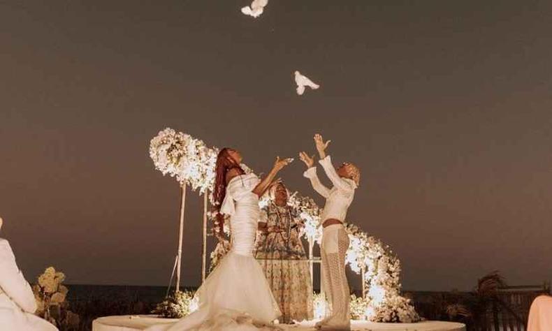 Majur e Josu esto no altar soltando pombas brancas. Ela  uma cantora trans no-binria preta com longas tranas ruivas e usa um vestido branco. Ele,  um homem preto de cabelos curtos e loiros presos em tranas e usa roupas brancas.