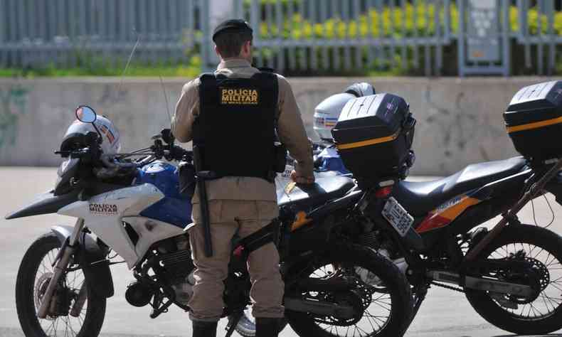 Na foto, policial fardado de costas em frente a duas motocicletas da PM