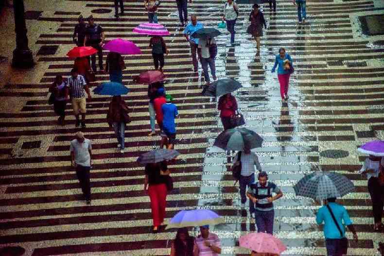 Pessoas caminhando em calada em dia chuvoso; algumas carregam guarda-chuva