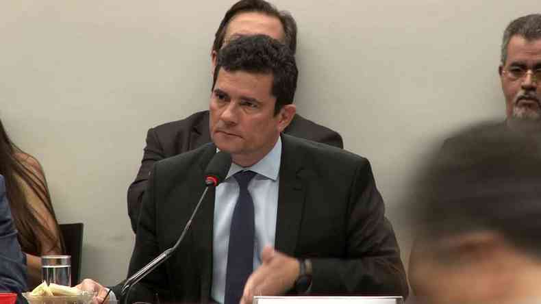 De terno e gravata escuros, sentado à mesa em sala de trabalho, o juiz Sergio Moro fala ao microfone 