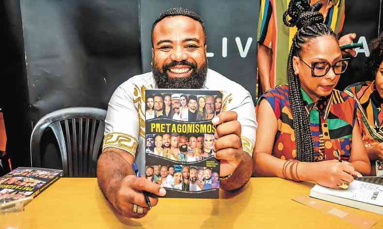 Sorrindo, sentado  mesa de autgrafos, Rodrigo Frana exibe a capa do livro 'pretagonismos' 