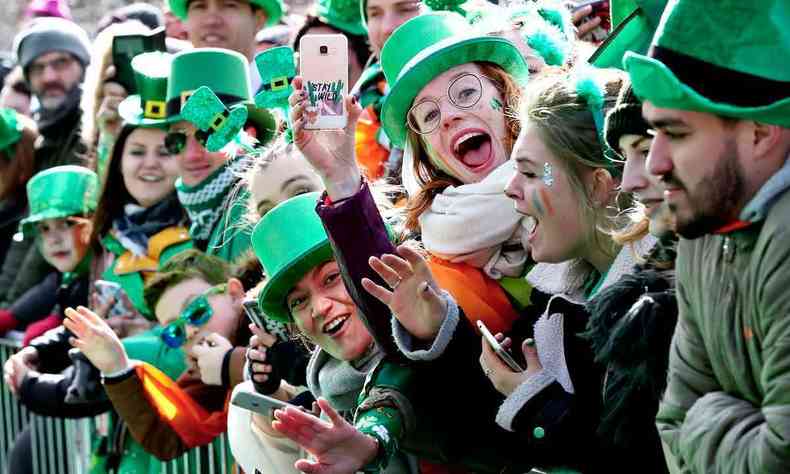 Pessoas com chapus e roupas verdes sorriem e mostram celulares, na festa St Patrick's Day 