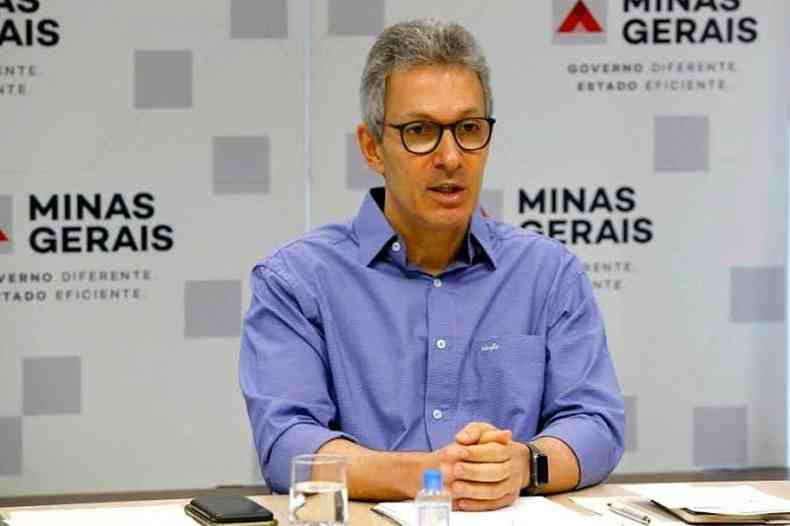 Romeu Zema sentado  mesa com uma painel do governo de Minas ao fundo