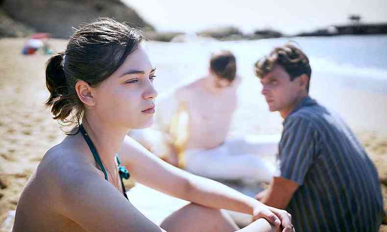 Garota com expresso sria, sentada na areia da praia, com dois rapazes ao fundo, em cena de o acontecimento
