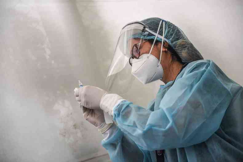 Profissional de sade com avental azul e mscara e toucas brancas, com uma seringa de vacina nas mos