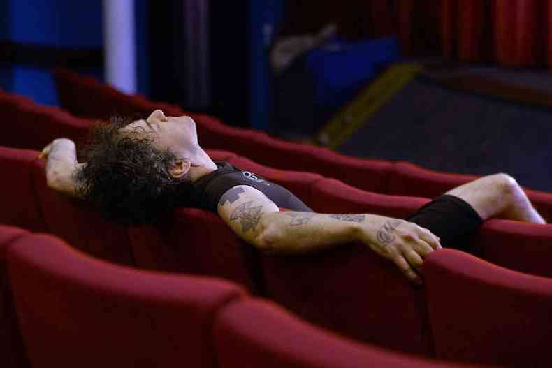 vestido de preto e com tatuagens na pele, homem abre os braos e recosta a cabea em poltronas vermelhas de uma sala de cinema vazia 