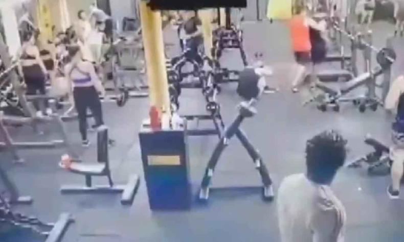 Academia com diversos aparelhos e pessoas. Vtima esta no ch de preto e agressor est sendo afastado por uma pessoa de vermelho