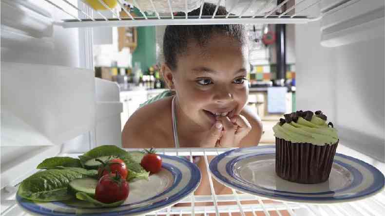 Menina olhando dentro da geladeira, onde est um prato de salada e um prato com cupcake
