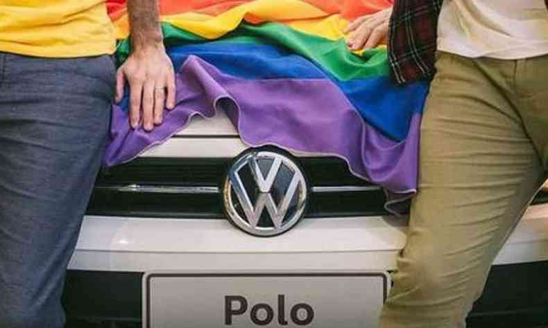 Frente do carro Polo, da Volkswagen, com a bandeira LGBT cobrindo o capô com casal gay, um de cada lado do carro