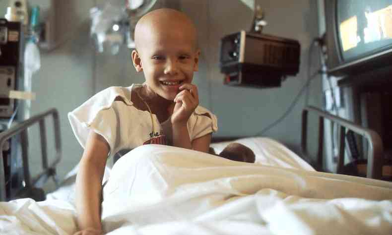 criança sorrindo em uma cama do hospital, já com queda de cabelo por causa do câncer