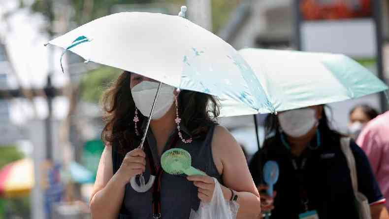 Duas mulheres usando ventiladores portteis e guarda-chuvas enquanto caminham na rua