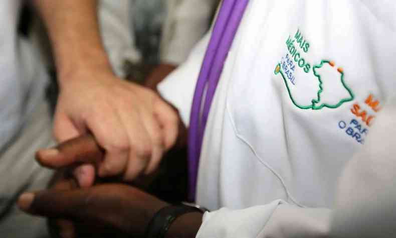 Uma pessoa negra com jaleco do programa Mais Mdicos segura as mos de um paciente branco