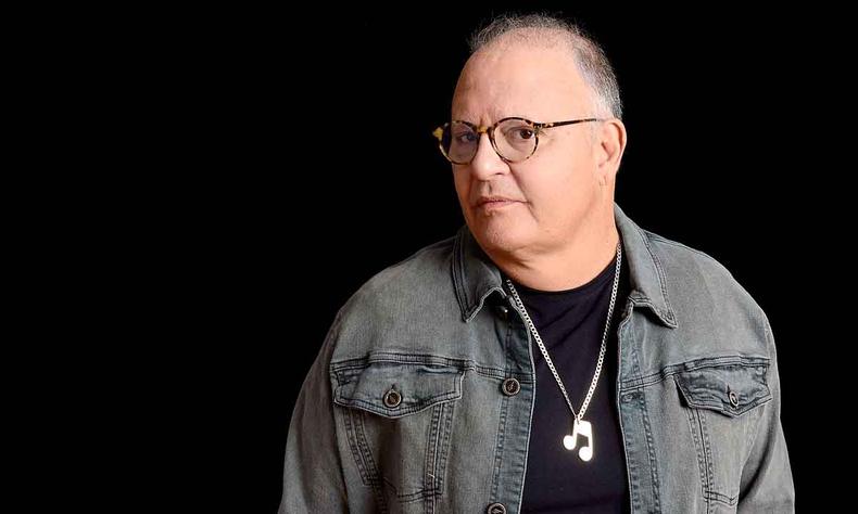 De jaqueta cinza, camisa preta e colar com pingente de nota musical, Guilherme Arantes olha de perfil para a cmera