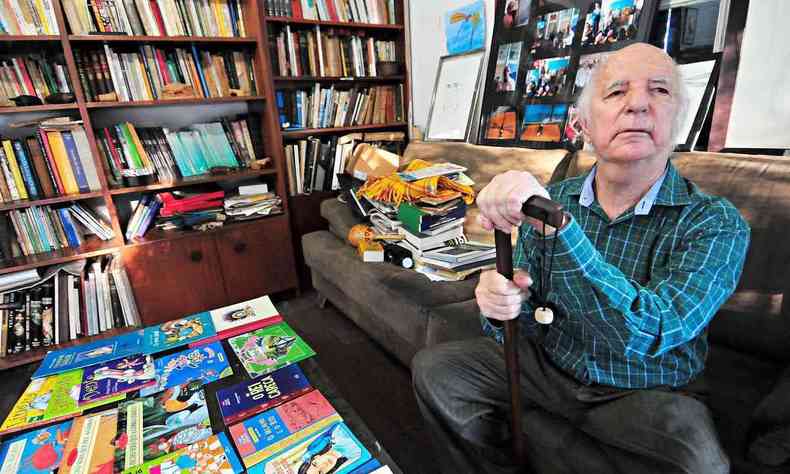 O escritor ngelo Machado olha para a cmera, em sua biblioteca, com seus livros infantis na mesa na frente dele, e tendo ao fundo uma estante