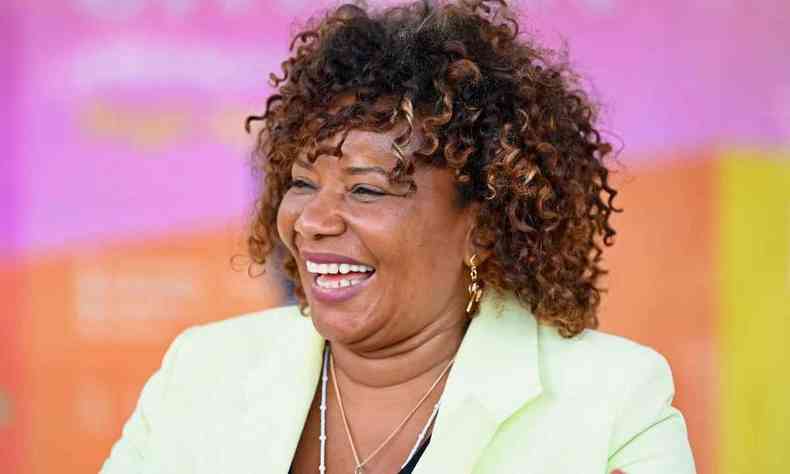 Margareth Menezes sorrindo: Mulher negra de cabelos cacheados castanhos, usando um blazer branco