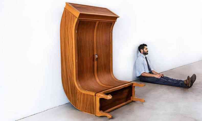 Escultor Francisco Nuk est sentado ao lado de um armrio que criou, que tambm est sentado no cho