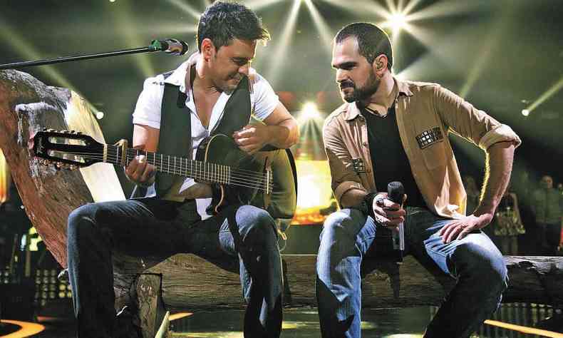 Zezé di Camargo (com violão) e Luciano (segurando microfone) se olham, sentados na beirada do palco 