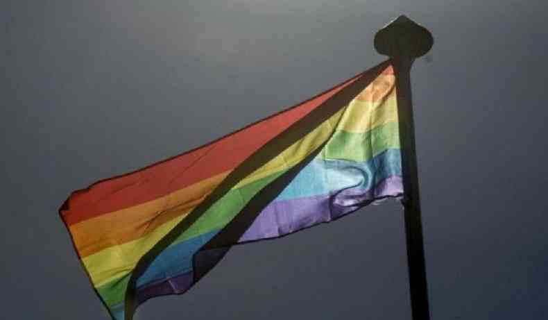 Bandeira com as cores que representam as cores da comunidade LGBTQIA+ hasteada em um mastro. No fundo da foto, um cu cinzento