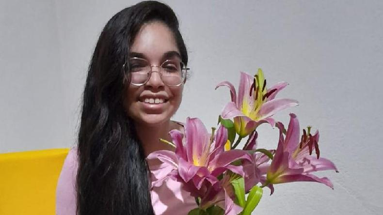 Rute atualmente, com 16 anos, segurando uma flor e sorrindo para a foto. Ela tem cabelos longos pretos e usa culos de grau