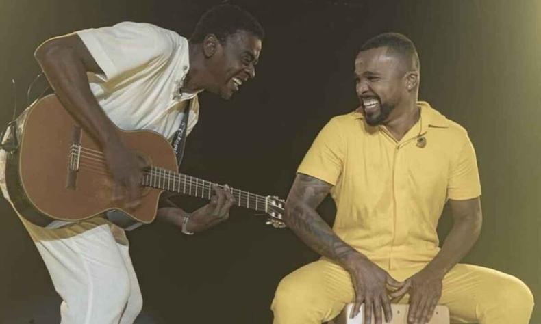 Seu Jorge e Alexandre sentados no palco, tocando violo e cantando, com luz azul ao alto 