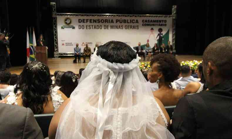 Mulher de costas com vu de noiva em frente ao palco, ao fundo um painel: 'Defensoria Pblica do Estado de Minas Gerais'