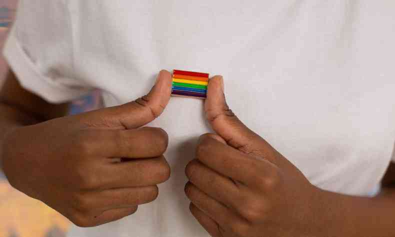 Foto mostra mãos de uma pessoa negra segurando um broche da bandeira LGBT que está em uma blusa branca