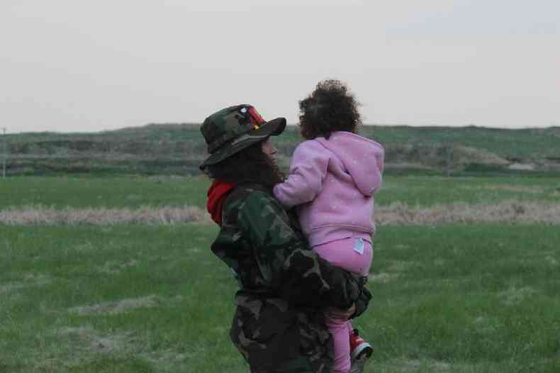 Cambria Harris com filha pequena no colo (de costas), em rea de gramado con su hija en brazos.