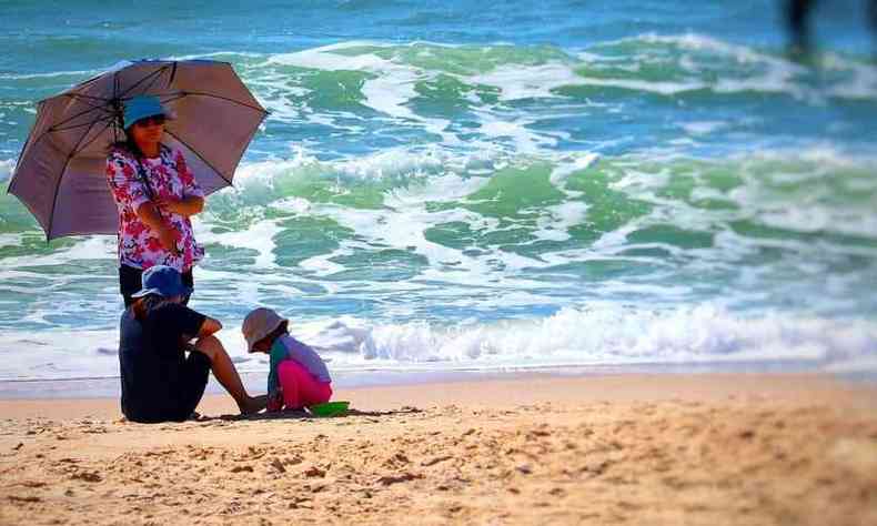 mulheres observam uma criana brincando na praia, quando todos esto de chapu, roupas e guarda-sol se protegendo dos raios solares