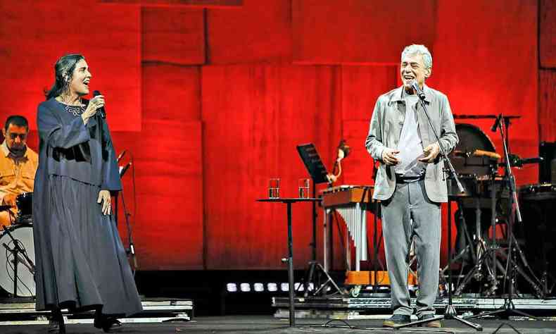 Mnica Salmaso (vestida de preto) e Chico Buarque (vestido de cinza), de p, lado a lado no palco, cantam e se olham, com cenrio de fundo em tom de vermelho