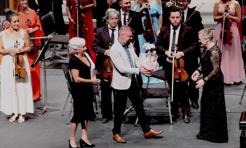 Claudia Malta observa Lenidas Oliveira entregando flores para a maestrina Ligia Amadio. Eles esto no palco do Palcio das Artes, com integrantes da Sinfnica ao fundo