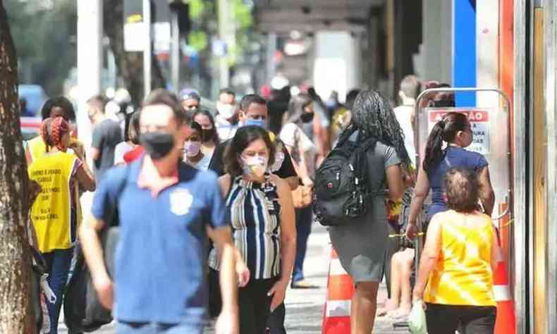 Pessoas, usando mscara, andando na regio central de BH. Na foto,  possvel ver uma grande quantidade de gente, mas somente duas esto viradas para cmera. Sendo eles: um homem com uniforme azul e mscara da cor preta; e uma senhora com uma blusa listrada e mscara da cor branca. 