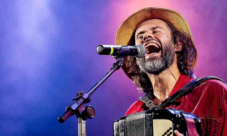 Com camisa vermelha, chapu de palha, Marcelo Jeneci toca sanfona e canta em frente a microfone no palco