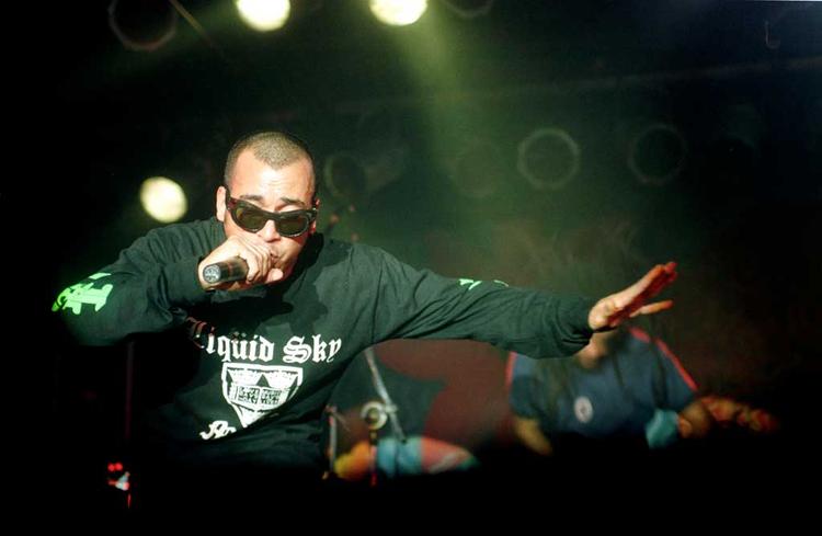Chico Science segura o microfone e canta durante show, estendendo a mo esquerda para a frente