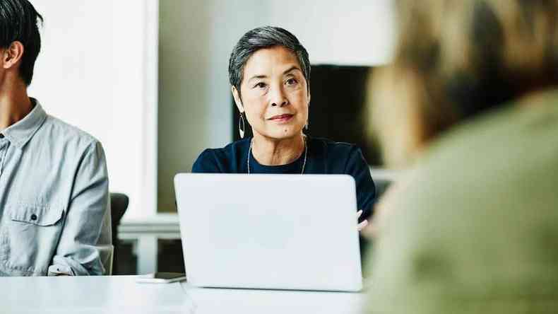 Mulher amarela em frente a um laptop conversa com algum quer est de costas