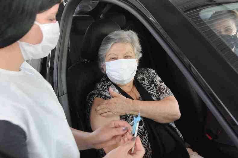 Idosa recebe vacina de dentro do carro, enfermeira em primeiro plano segurando seringa
