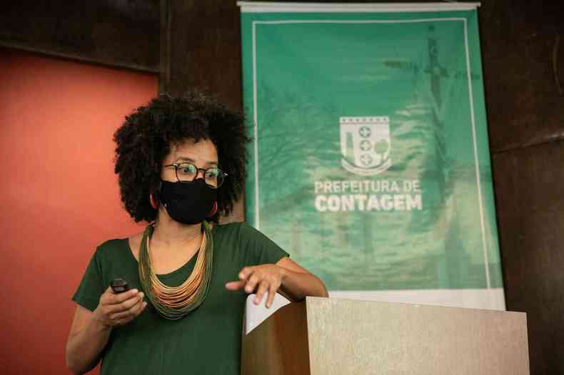 Lorena Luiza veste roupa verde, usa mscara e fala em pblico. Ao fundo, painel da Prefeitura Municipal de Contagem