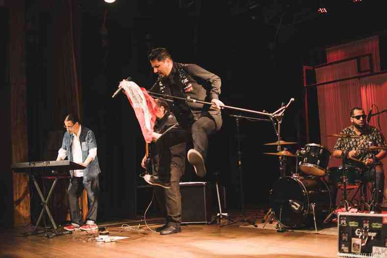 Saulo Wolf and the Gang, vestido de preto, ergue a perna e segura microfone em palco 