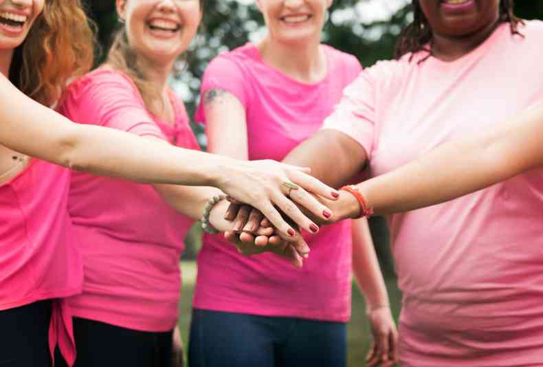 Cinco mulheres, vestindo camisa rosa, juntam as mos sorrindo, em gesto de cooperao e unio contra o cncer de mama, demonstrando a necessidade de divulgar informaes com a campanha 'Outubro Rosa' para conhecer mais sobre a condio e a importncia da mamografia para um diagnstico precoce 