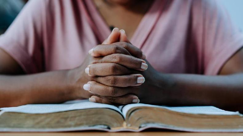 Fotografia mostra mos de uma mulher negra entrelaadas sobre um livro como quem faz uma orao