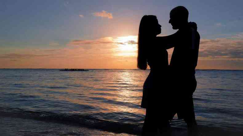 Foto de casal abraçado na praia diante do pôr do sol