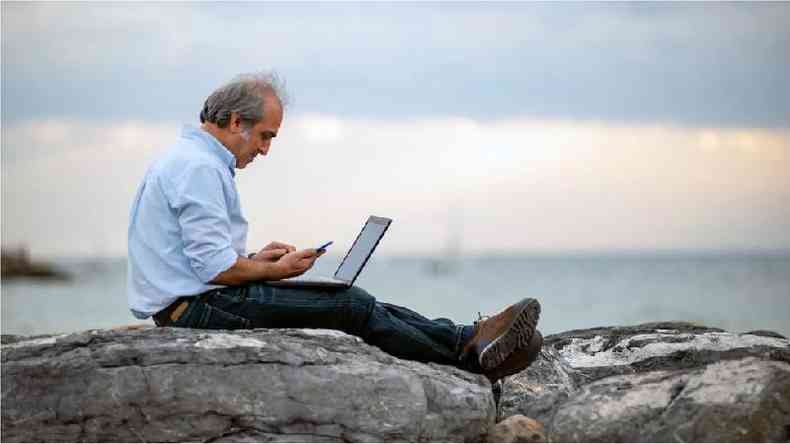 Homem branco de cabelo grisalho sentado em uma rocha com um laptop no colo